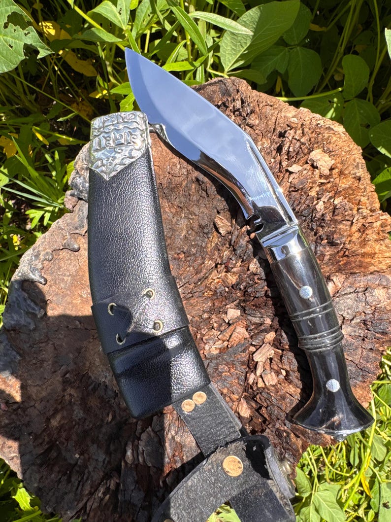 May include: A polished steel kukri knife with a curved blade and a black handle, resting on a wooden surface. The knife is accompanied by a black leather sheath with decorative metal accents and a belt loop. The blade reflects the surrounding environment.