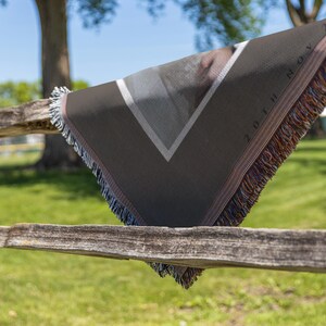 May include: A dark gray woven throw blanket with a geometric design and fringe. The blanket has a white and gray triangle pattern and the text "20TH NOV". The blanket is draped over a wooden fence.