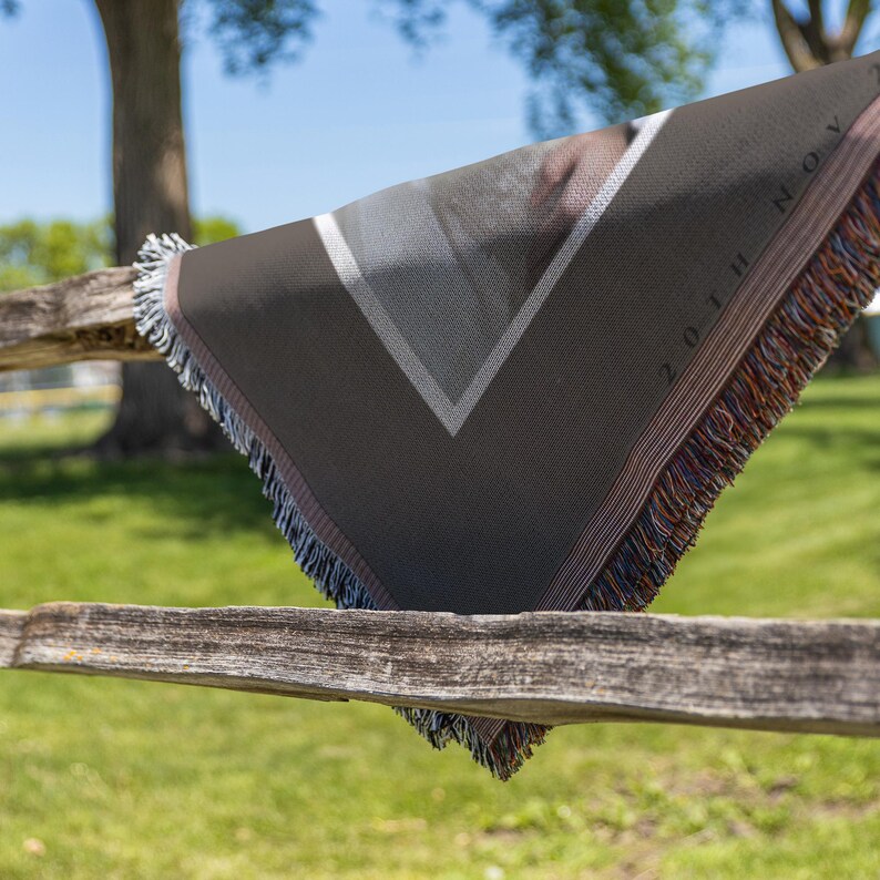 May include: A dark gray woven throw blanket with a geometric design and fringe. The blanket has a white and gray triangle pattern and the text "20TH NOV". The blanket is draped over a wooden fence.