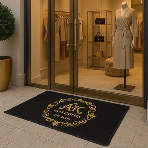 May include: Black rectangular doormat with a gold floral design and the text "AK Anna Katrina Est 2000". The doormat is placed in front of a glass door entrance to a boutique.