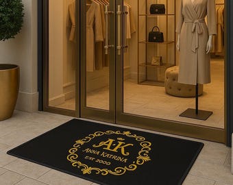 Puede incluir: Felpudo rectangular negro con un diseño floral dorado y el texto "AK Anna Katrina Est 2000". El felpudo está colocado frente a la puerta de entrada de cristal de una boutique.
