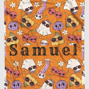 May include: Orange blanket with a repeating pattern of cartoon skulls, cherries, ghosts, and disco balls, all wearing sunglasses. The word "SAMUEL" is printed in large black letters across the center.