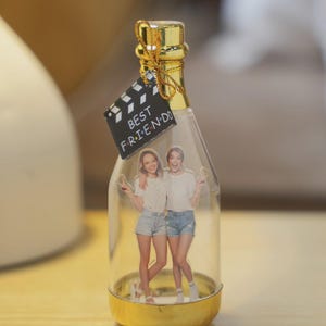 Puede incluir: Una botella de vidrio transparente con base y tapa doradas, que contiene una foto de dos mujeres. Una etiqueta negra con la inscripción "BEST FRIEND" está adjunta. Objeto decorativo y de recuerdo.