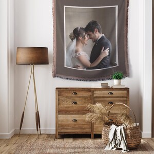 May include: A woven tapestry featuring a framed wedding photo of a couple embracing. The tapestry is brown with a white border and fringed edges. It hangs above a wooden dresser with a basket and a floor lamp.