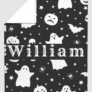May include: A black and white Halloween-themed blanket featuring a pattern of ghosts, jack-o'-lanterns, stars, and spiders. The name "William" is prominently displayed in white. The blanket is folded, revealing a white underside.