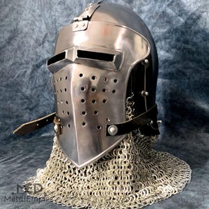 Puede incluir: Un casco medieval de acero pulido con visera con ranuras para los ojos y orificios de ventilación. El casco incluye una protección para el cuello de cota de malla y correas de cuero. El diseño recuerda a las armaduras históricas.