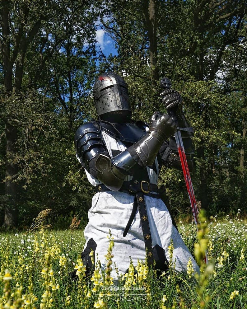 May include: A knight in full armor kneels in a field of yellow flowers, holding a sword. The armor is black and silver, with a helmet featuring a red cross. The sword has a red blade. The knight is wearing a white tunic with a black belt.