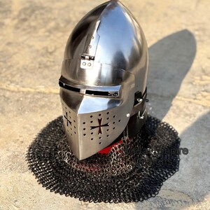 May include: A polished steel knight's helmet with a cross design on the faceplate and a chainmail neck guard. The helmet has a rounded top and a visor. The chainmail is black and surrounds the base of the helmet.