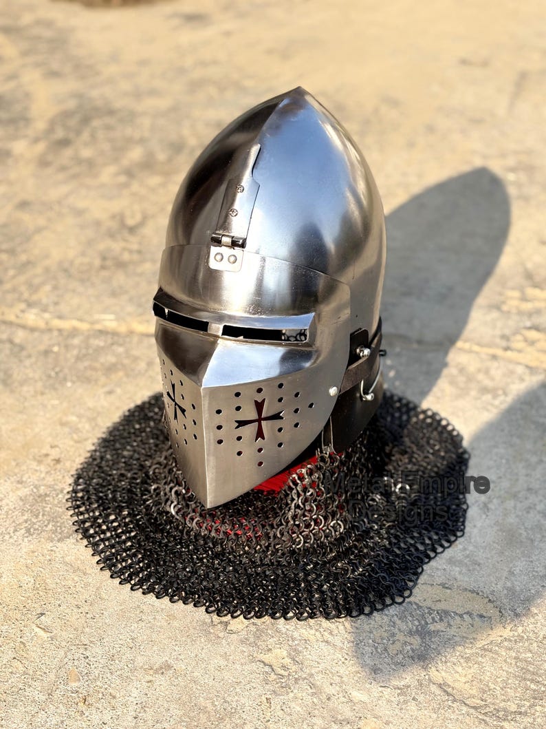 May include: A polished steel knight's helmet with a cross design on the faceplate and a chainmail neck guard. The helmet has a rounded top and a visor. The chainmail is black and surrounds the base of the helmet.