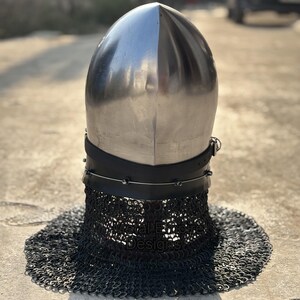 May include: A polished silver medieval helmet with a rounded top and a black leather neck guard. The helmet features a chainmail aventail for neck protection. The helmet is designed for historical reenactment or display.