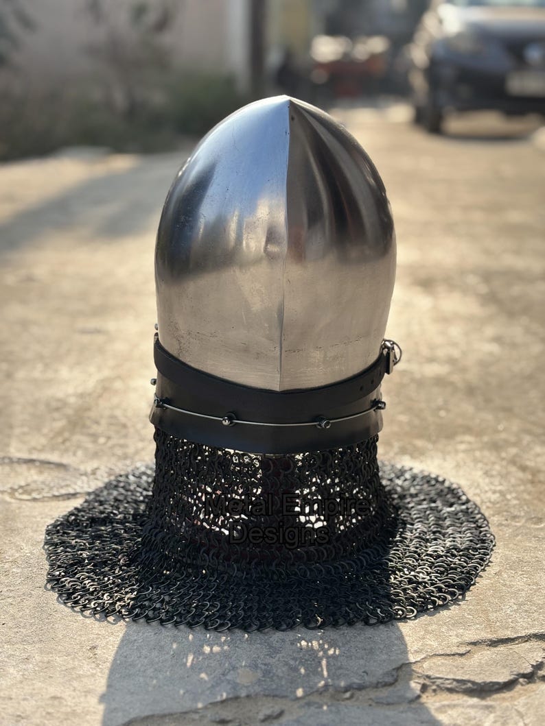 May include: A polished silver medieval helmet with a rounded top and a black leather neck guard. The helmet features a chainmail aventail for neck protection. The helmet is designed for historical reenactment or display.