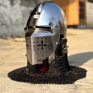 May include: A polished steel knight's helmet with a visor featuring a cross design and ventilation holes. The helmet is connected to a black chainmail coif. The helmet is a historical replica.