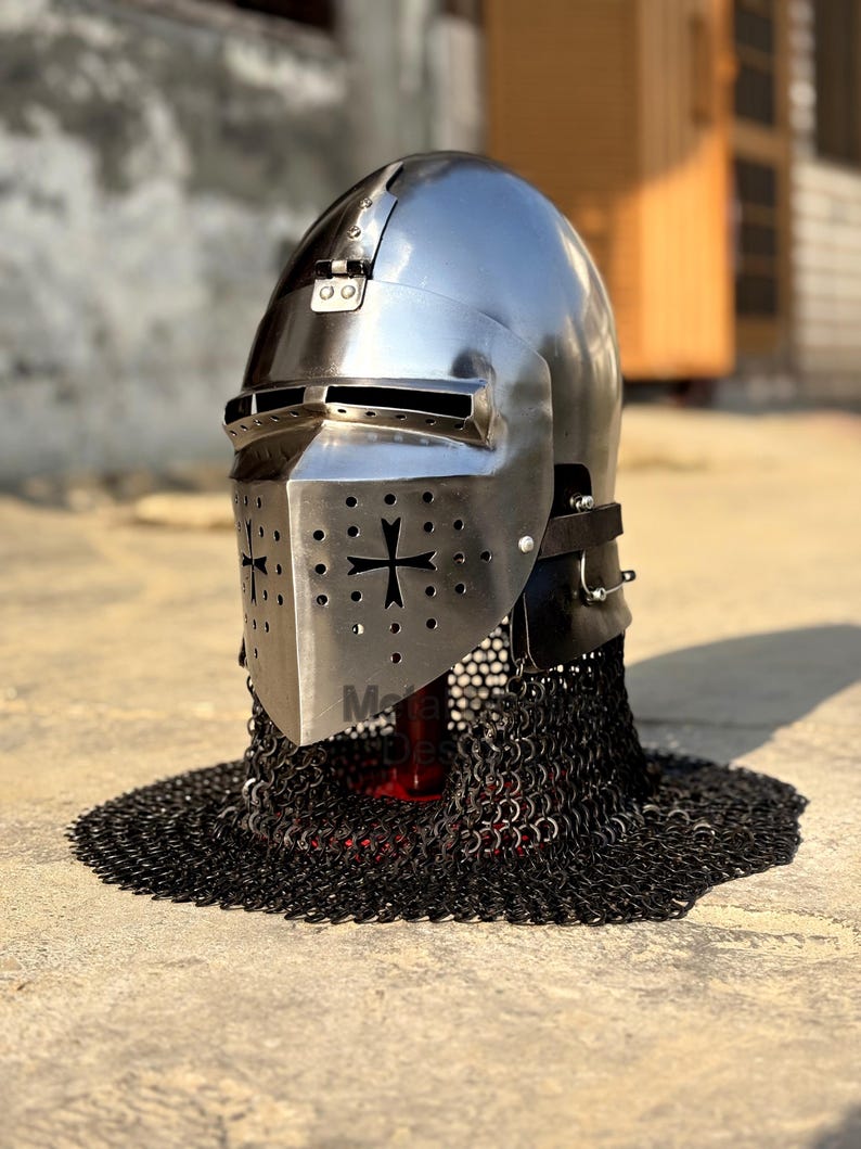 May include: A polished steel knight's helmet with a visor featuring a cross design and ventilation holes. The helmet is connected to a black chainmail coif. The helmet is a historical replica.