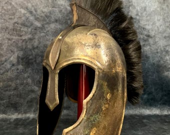 May include: A bronze-colored, antique-style helmet with a dark, feathery plume. The helmet has a curved design with a black visor and is displayed on a red stand. The helmet's surface shows signs of wear, giving it a vintage appearance.