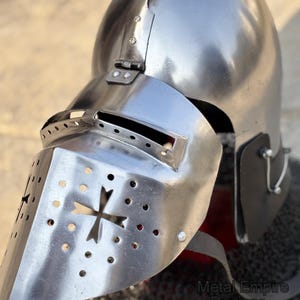 May include: A polished silver medieval helmet with a visor featuring a cross design and ventilation holes. The helmet has a rounded top and is partially covering a chainmail coif. The text "Metal Empire Designs" is visible.