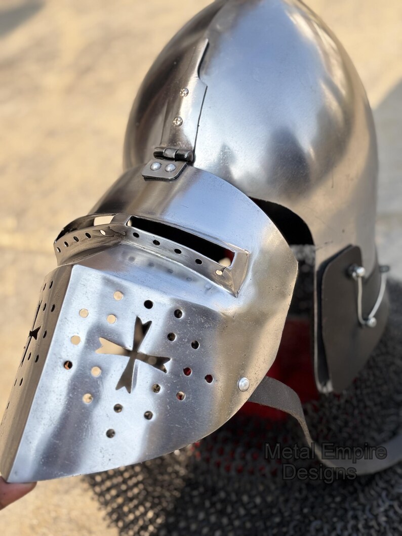May include: A polished silver medieval helmet with a visor featuring a cross design and ventilation holes. The helmet has a rounded top and is partially covering a chainmail coif. The text "Metal Empire Designs" is visible.