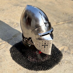 May include: A polished steel medieval helmet with a visor and a cross emblem. The helmet is set on a black chainmail coif. The helmet has a rounded top and a hinged visor with small eye slits.