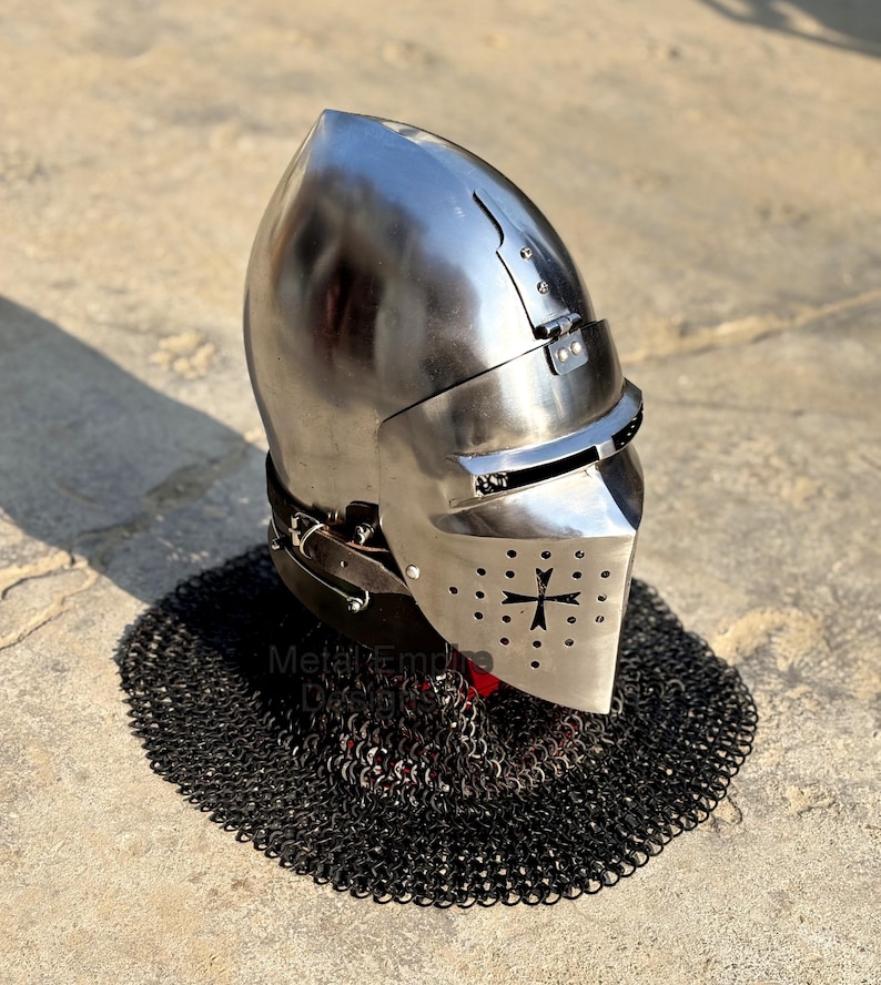 May include: A polished steel medieval helmet with a visor and a cross emblem. The helmet is set on a black chainmail coif. The helmet has a rounded top and a hinged visor with small eye slits.