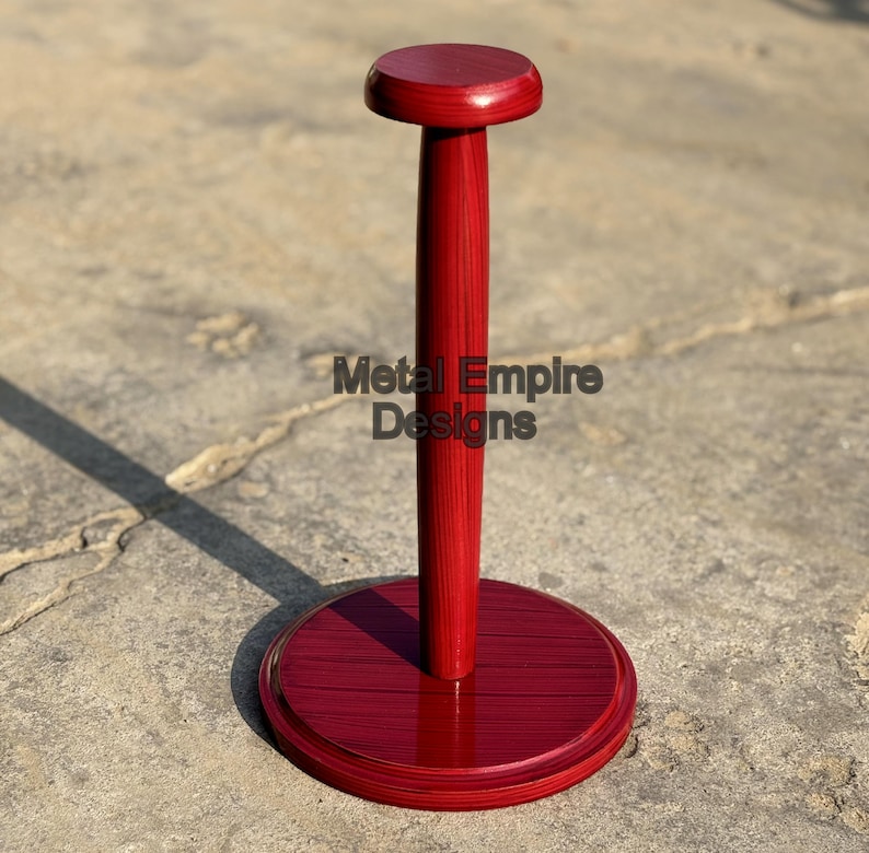 May include: A red wooden helmet stand with a round base and top, and a central pole. The stand has the text "Metal Empire Designs" on the pole. The stand is designed to hold a helmet.