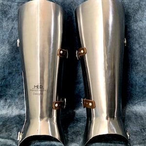 May include: A pair of polished steel greaves, part of medieval armor. Each greave features a curved design, leather straps, and metal buckles. The product is from MetalEmpire Designs.