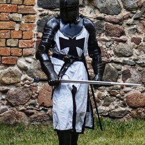 May include: A person in full medieval armor, including a helmet, black and white tunic with a cross, and gauntlets, stands holding a sword. The armor is detailed with black metal plates. The background is a stone brick wall and green grass.