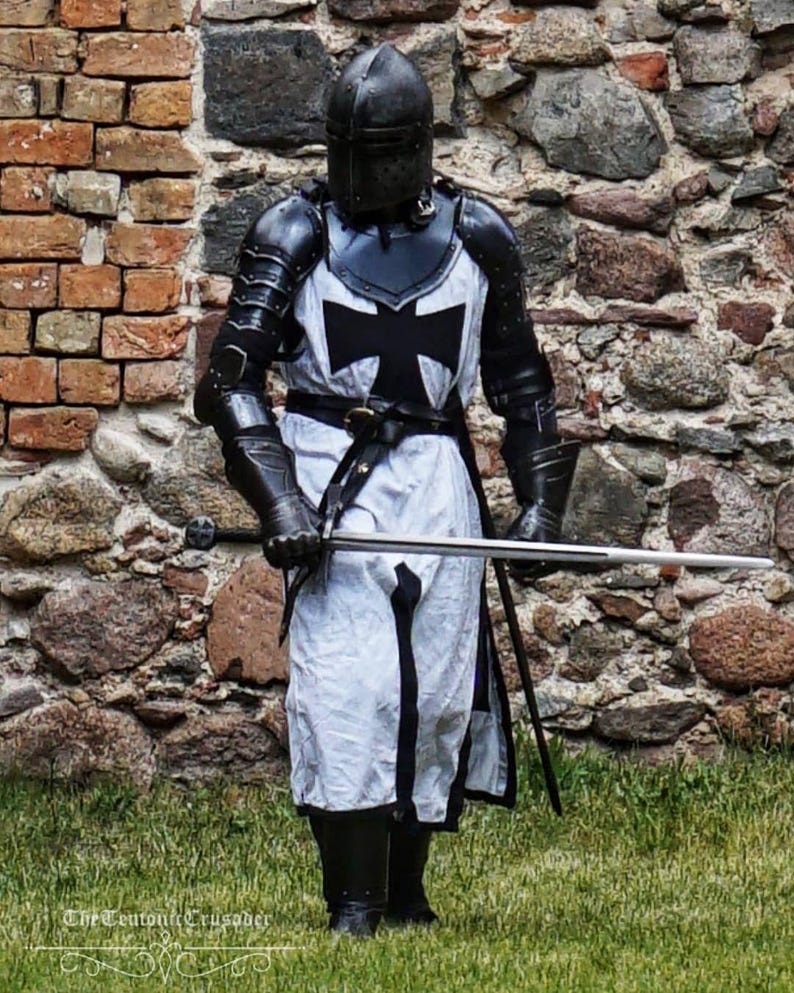 May include: A person in full medieval armor, including a helmet, black and white tunic with a cross, and gauntlets, stands holding a sword. The armor is detailed with black metal plates. The background is a stone brick wall and green grass.