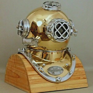 May include: A vintage brass diving helmet with silver accents, mounted on a wooden base. The helmet features portholes, a top vent, and a circular faceplate. The base has a semi-circular shape. The helmet has a reflective surface.