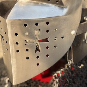 May include: Close-up of a polished metal knight's helmet. The helmet features a cross-shaped cutout and numerous small circular ventilation holes. The text "Metal Empire Designs" is visible on the lower right. The helmet is part of a medieval armor collection.