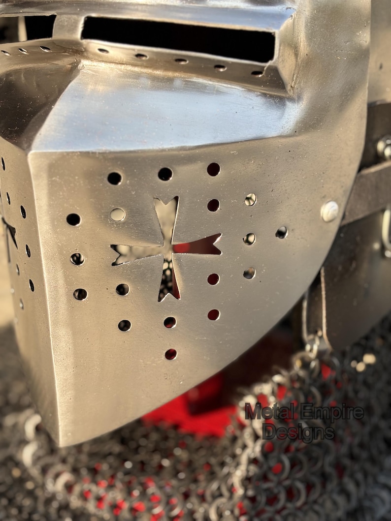 May include: Close-up of a polished metal knight's helmet. The helmet features a cross-shaped cutout and numerous small circular ventilation holes. The text "Metal Empire Designs" is visible on the lower right. The helmet is part of a medieval armor collection.