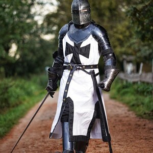 May include: A full suit of medieval armor, including a helmet, chest plate, and leg guards. The armor is primarily black and silver, with a white tunic featuring a large black cross. The knight is holding a sword and walking on a dirt path.
