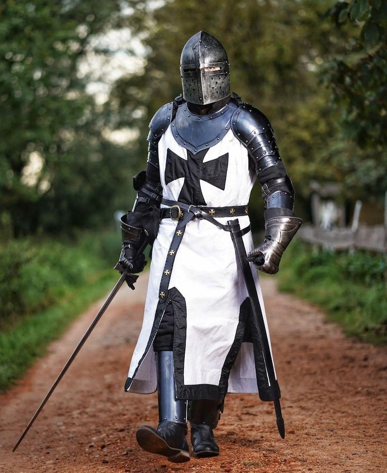 May include: A full suit of medieval armor, including a helmet, chest plate, and leg guards. The armor is primarily black and silver, with a white tunic featuring a large black cross. The knight is holding a sword and walking on a dirt path.