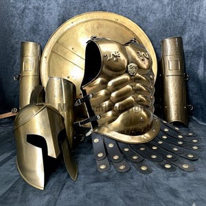 Könnte beinhalten: Ein komplettes Set antiker messingfarbener Rüstung, einschließlich Helm, Brustplatte, Schild sowie Arm- und Beinschützern. Die Rüstung weist aufwendige Details und ein verwittertes Finish auf.