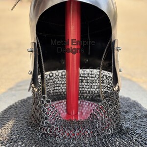 May include: A medieval-style helmet with a polished metal finish, featuring a chainmail neck guard. The helmet has a dark interior and is supported by a red wooden pole. The text "Metal Empire Designs" is visible on the helmet.