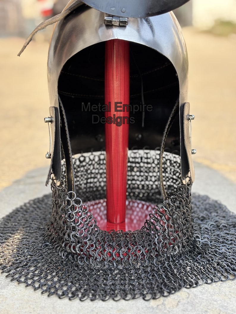 May include: A medieval-style helmet with a polished metal finish, featuring a chainmail neck guard. The helmet has a dark interior and is supported by a red wooden pole. The text "Metal Empire Designs" is visible on the helmet.