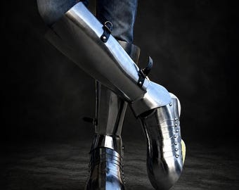 May include: Polished steel leg armor, featuring articulated plates and straps. The armor is worn over blue jeans, with a dark background. The metal reflects light, highlighting the craftsmanship and historical design.