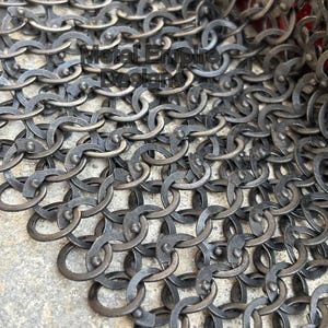 May include: Close-up of a section of chainmail armor. The metal rings are interlocked, creating a dense, flexible fabric. The armor has a dark, aged appearance, with visible wear and tear. The text "Metal Empire Designs" is visible.