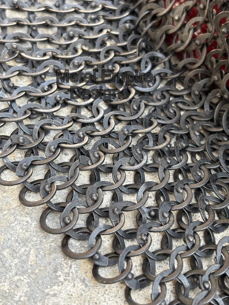 May include: Close-up of a section of chainmail armor. The metal rings are interlocked, creating a dense, flexible fabric. The armor has a dark, aged appearance, with visible wear and tear. The text "Metal Empire Designs" is visible.