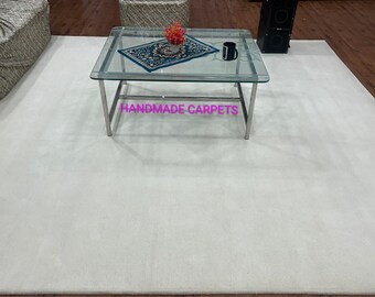 Puede incluir: Una gran alfombra blanquecina domina la escena, con una mesa de centro con tapa de cristal en el centro. La mesa tiene una estructura de metal plateado y una pequeña alfombra estampada encima. Una taza negra y un pequeño arreglo floral están sobre la mesa. El texto "HANDMADE CARPETS" es visible.