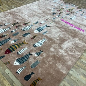 Könnte beinhalten: Ein rechteckiger, brauner Teppich mit einem Muster aus stilisierten Fischen in verschiedenen Farben, darunter Weiß, Schwarz und Gold. Der Teppich trägt den Text "HANDMADE CARPETS" in Pink. Die Fische sind in einem fließenden, aquatischen Design angeordnet.