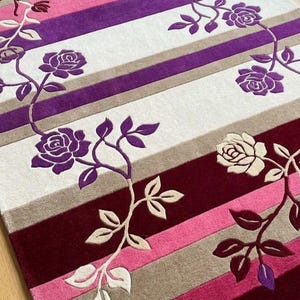Könnte beinhalten: Ein rechteckiger Teppich mit horizontalen Streifen in den Farben Bordeaux, Pink, Violett und Beige. Der Teppich ist mit einem sich wiederholenden Muster aus kletternden Rosenranken mit dunkelvioletten und cremefarbenen Blüten und grünen Blättern verziert.