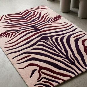 Könnte beinhalten: Ein rechteckiger Teppich mit hellrosa Grundfarbe und einem sich wiederholenden, symmetrischen Muster aus dunkelvioletten und tiefroten, zebraartigen Streifen, die sich über die gesamte Oberfläche des Textilbodens erstrecken.