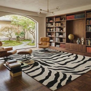 Könnte beinhalten: Ein Wohnzimmer mit einem zebra gemusterten Teppich, zwei braunen Ledersesseln und einem passenden Hocker. Ein dunkles Holz Bücherregal ist mit Büchern gefüllt. Ein Fenster blickt auf einen Garten mit Bäumen und Grün. Ein kleiner schwarzer Tisch hält Bücher und eine dekorative Schale.