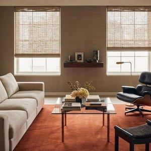 Könnte beinhalten: Ein modernes Wohnzimmer mit beigem Sofa, Glastisch und schwarzem Ledersessel. Der Raum verfügt über einen rostfarbenen Teppich, gewebte Fensterrollos und ein minimalistisches Design. Ein kleines Holzregal zeigt dekorative Gegenstände.