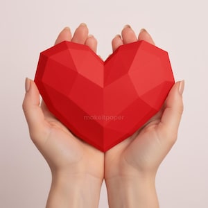 Peut inclure: Un cœur rouge vif, facetté, est tenu dans des mains jointes. Le cœur a un design géométrique et le texte "makeitpaper" est imprimé en bas à droite. Le fond est d'une couleur douce et neutre.