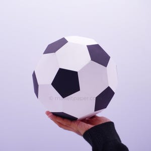 Op de afbeelding: Een papieren voetbalmodel met een klassiek geometrisch patroon van zwarte vijfhoeken en witte zeshoeken. Het oppervlak toont de tekst makeitpaper.com in kleine, lichtgrijze letters op een van de witte panelen van de bol.