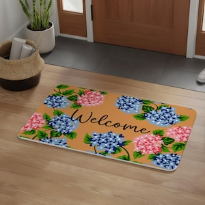 Puede incluir: Una alfombra rectangular con un fondo marrón y un diseño floral con hortensias azules y rosas. La palabra "Welcome" está escrita en escritura negra en el centro. La alfombra está colocada frente a una puerta de madera.