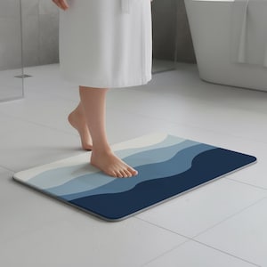 May include: A rectangular bath mat with a wave design in shades of blue and white. The mat has a gray border and is placed on a white tiled floor. The mat is approximately 24 inches long and 16 inches wide.