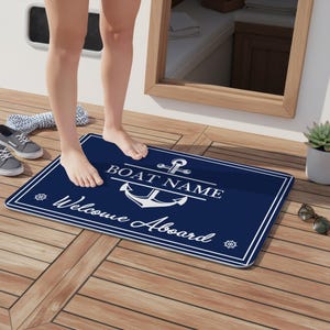May include: A navy blue rectangular welcome mat with a white border and nautical design. The mat features an anchor graphic and the words "BOAT NAME" and "Welcome Aboard" in white. The mat is on a wooden deck.