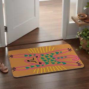 May include: Rectangular doormat with a whimsical design. The mat features a stylized animal figure in pink and teal, set against a tan background. Yellow lines radiate from the top of the figure. The mat is placed in a doorway.