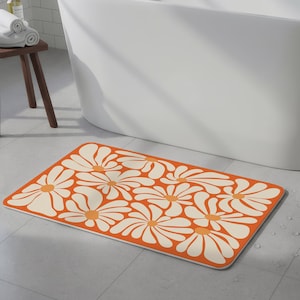 May include: An orange rectangular bath mat with a white daisy pattern. The mat is placed in front of a white bathtub. The daisy flowers have a yellow center and white petals. The mat is made of a soft, absorbent material.